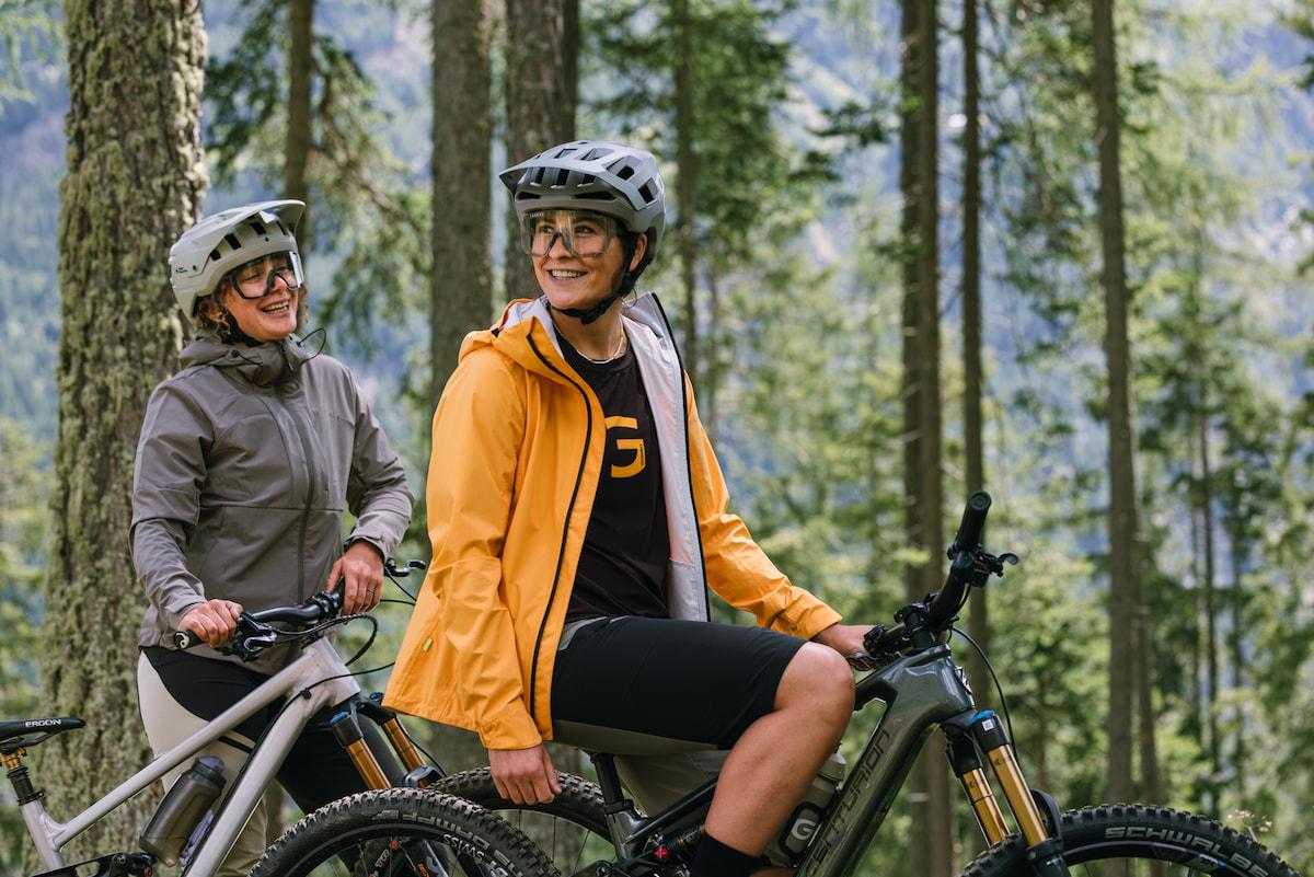 Fahrrad fahren ist sehr gelenkschonend und Gold wer bei Arthrose, nach Operationen oder bei Übergewicht. Zwei Mountainbikerinnen im Wald, die vordere sitzt auf dem Fahrrad und schaut nach hinten, die hintere steht neben dem Rad und lacht