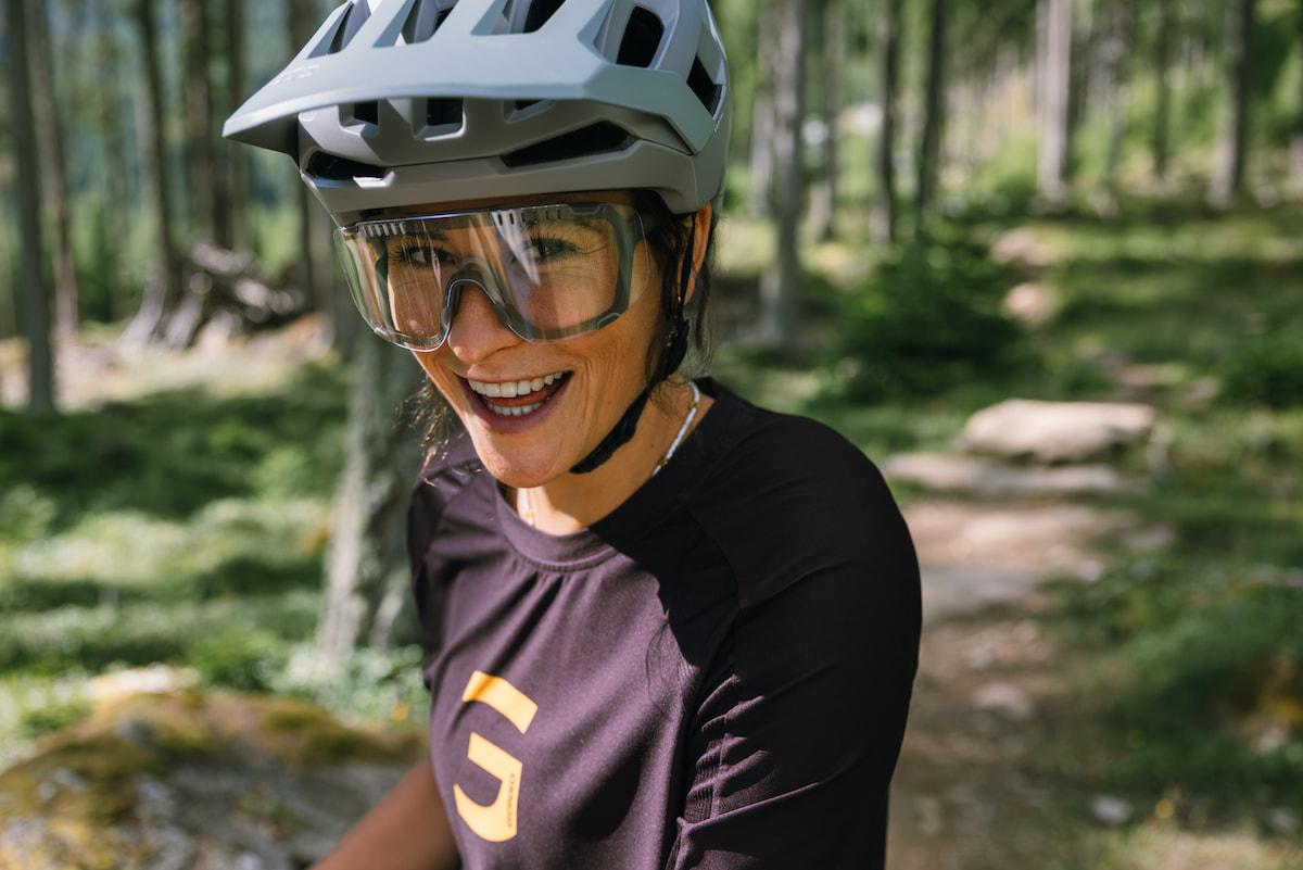 Ausdauerbewegung an der frischen Luft schüttet Endorphine aus, senkt Cortisolspiegel und verbessert die Schlafqualität. Frau lacht fröhlich in die Kamera, im Hintergrund ist ein Waldtrail zu sehen