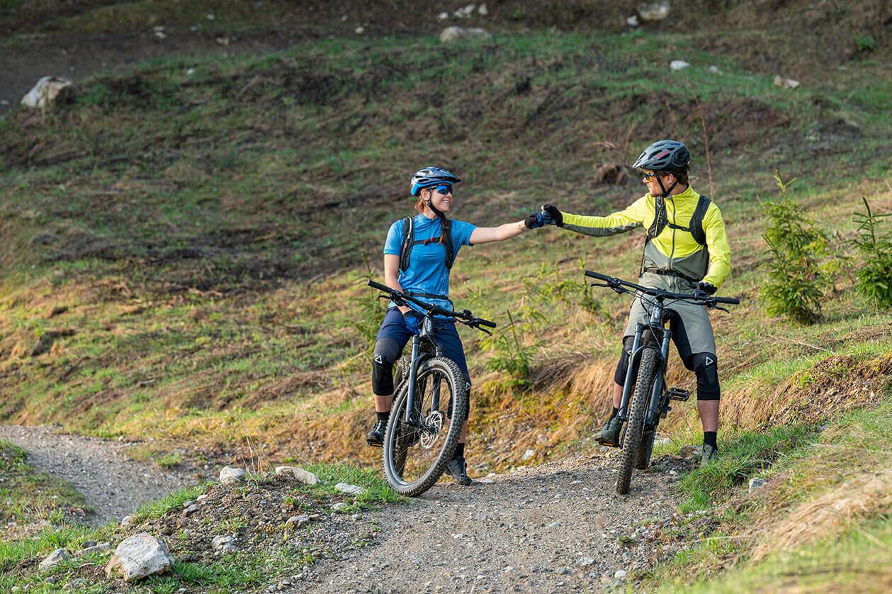 Bei einem kurzen Stop kann es helfen, die Arme hängen zu lassen, die Finger zu spreizen und zu schließen sowie die Handgelenke kreisen zu lassen. Zwei Fahrradfahrer machen auf Gravel Trail kurzen Stop, geben sich die Faust. Beide grinsen.
